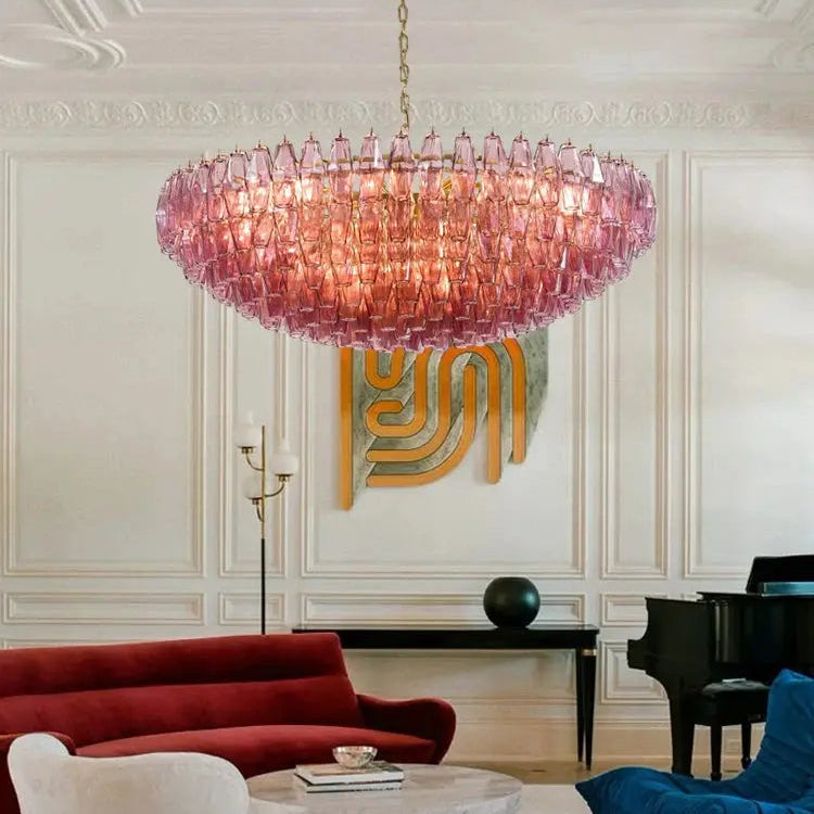 A large pink glass chandelier hangs in an elegant living room with cream paneled walls, a red sofa, a white armchair, a blue beanbag chair, and a marble coffee table.