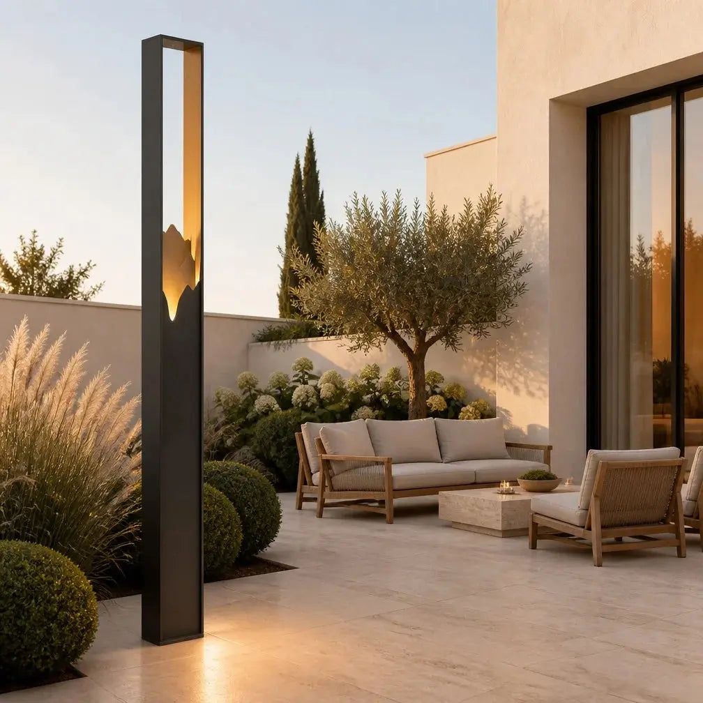 A modern tall rectangular outdoor LED light fixture with a warm glowing leaf-shaped interior, installed on a sunlit patio next to a contemporary lounge set and landscaped garden.