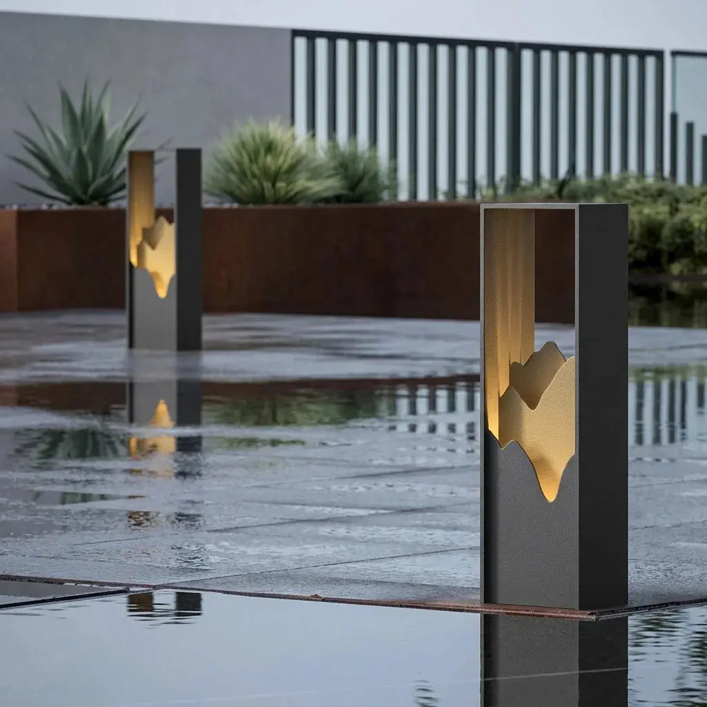Three modern rectangular bollard lights with a dark gray finish and a glowing gold abstract mountain cutout, set in a wet outdoor plaza with reflections.