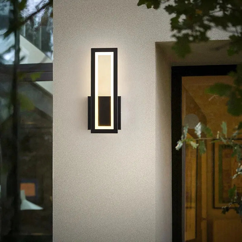 A modern rectangular wall sconce light fixture with a black frame and warm white LED lighting, mounted on a textured beige stucco exterior wall next to a glass door.
