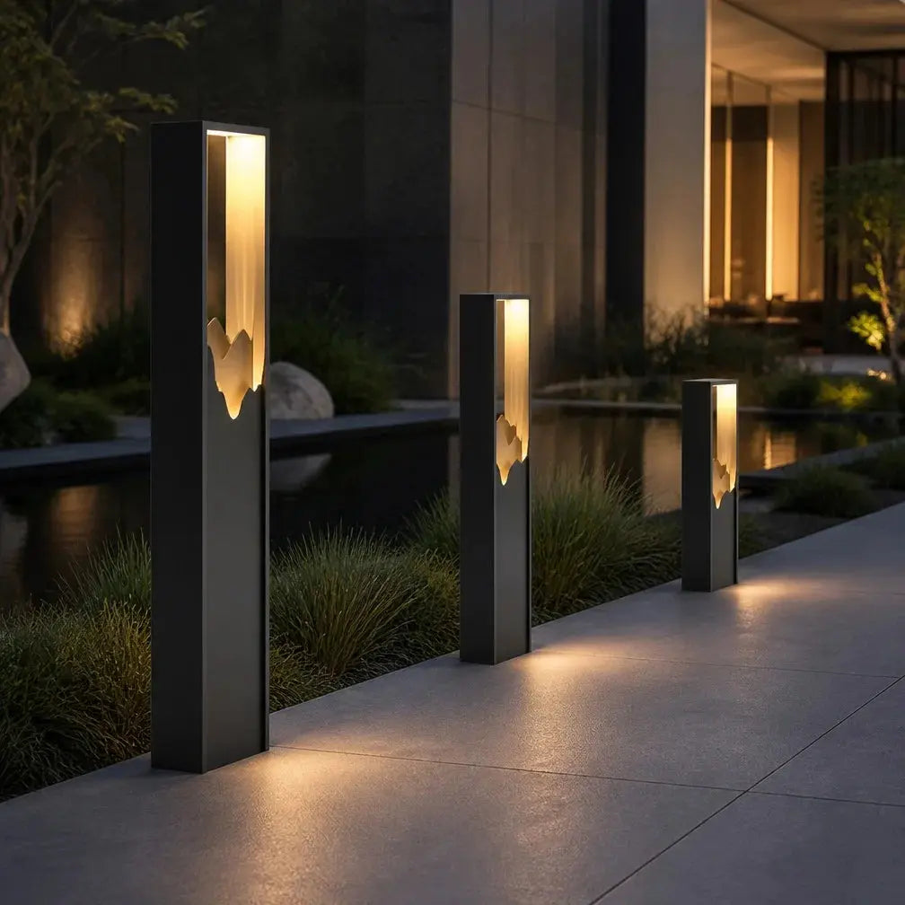 Three modern black rectangular outdoor bollard lights with glowing warm white geometric cutout panels, arranged along a paved walkway beside a reflective water feature at dusk.