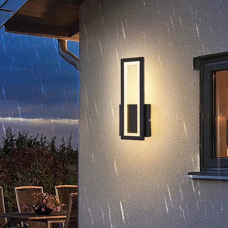 A modern vertical rectangular outdoor wall sconce mounted on a textured stucco house wall, glowing with warm light at dusk. The scene includes a window, outdoor dining furniture, and rain streaks visible in the air.